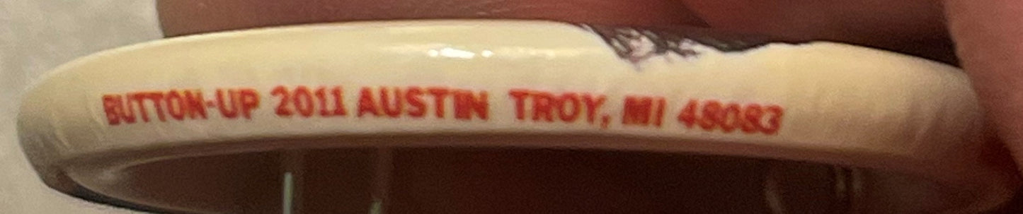 Close-up of curved white pin edge with red stamped text reading "BUTTON-UP 2011 AUSTIN TROY, ILL 48033