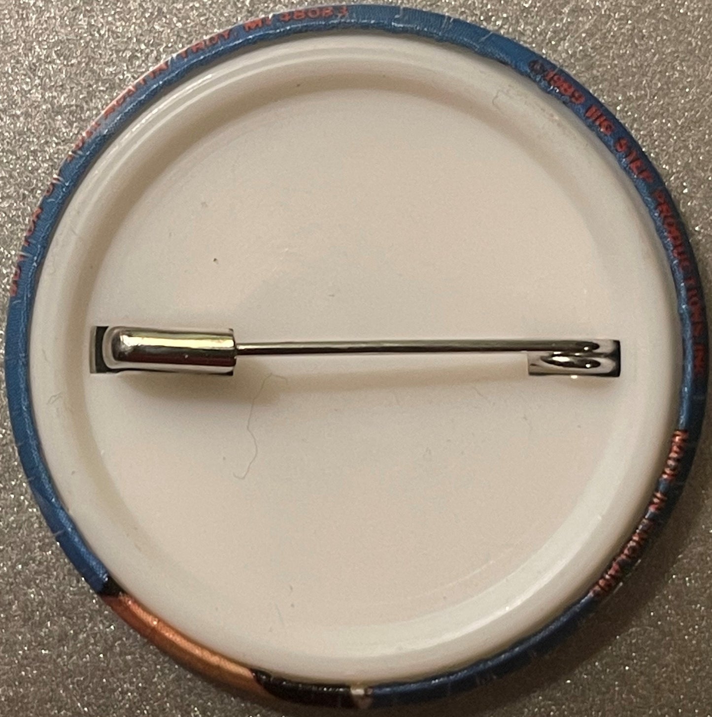Circular metal pin badge with blue and red fabric rim displayed on cream ceramic plate against concrete surface, shown from a