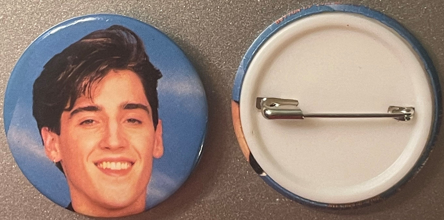 Front and back of a blue circular pin featuring a smiling man with dark hair against a blue background, with a silver pin cla