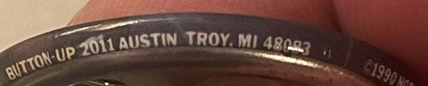 Close-up of embossed text on black metal pin rim reading "BUTTON-UP 2011 AUSTIN TROY, MI 48027