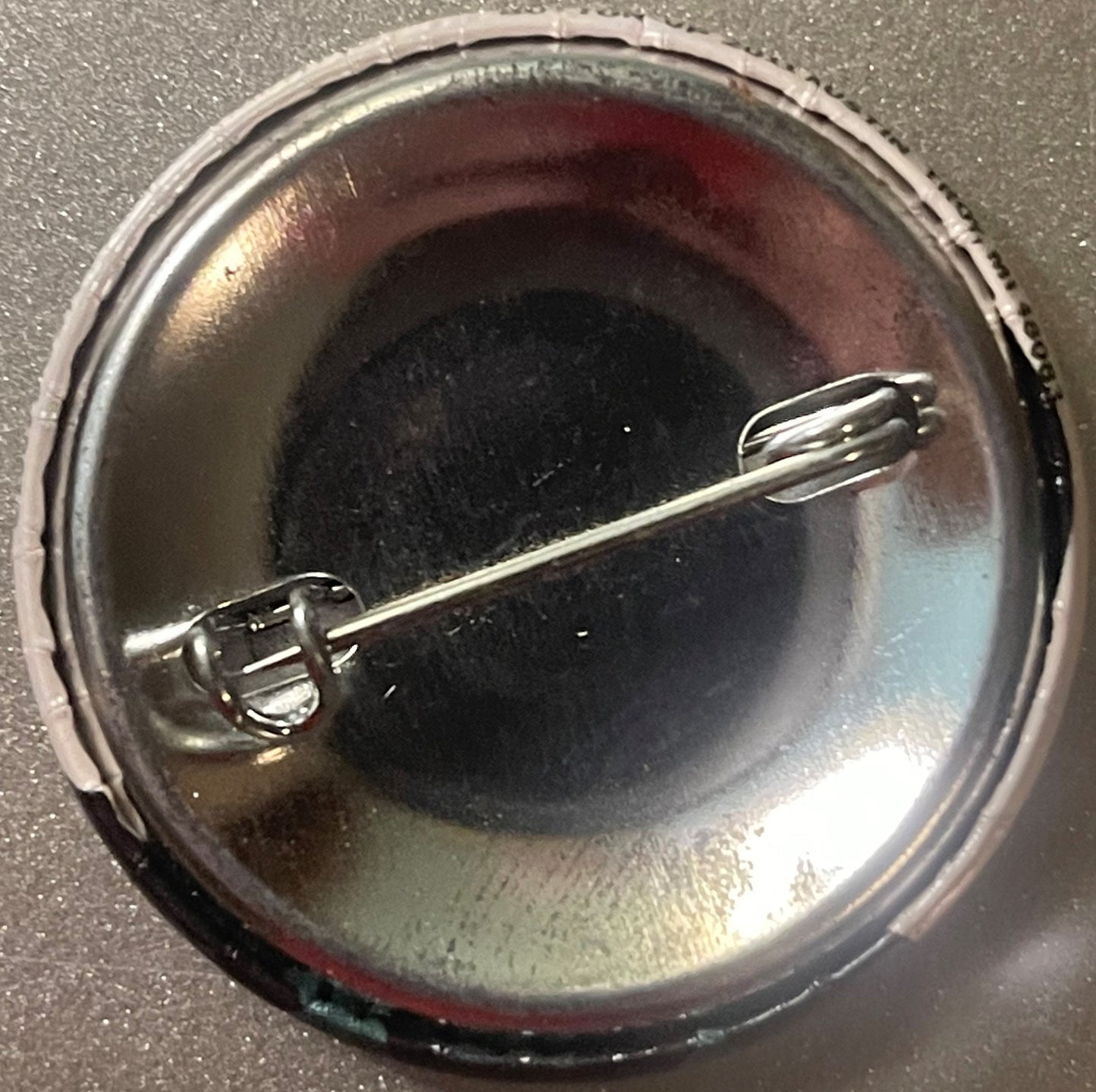 Circular metal pin with dark center displayed in clear plastic case, shown from above with backing card visible underneath