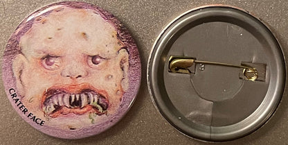 Vintage pinback button with grotesque face featuring purple-tinted skin, closed eyes, and prominent teeth, shown front and ba