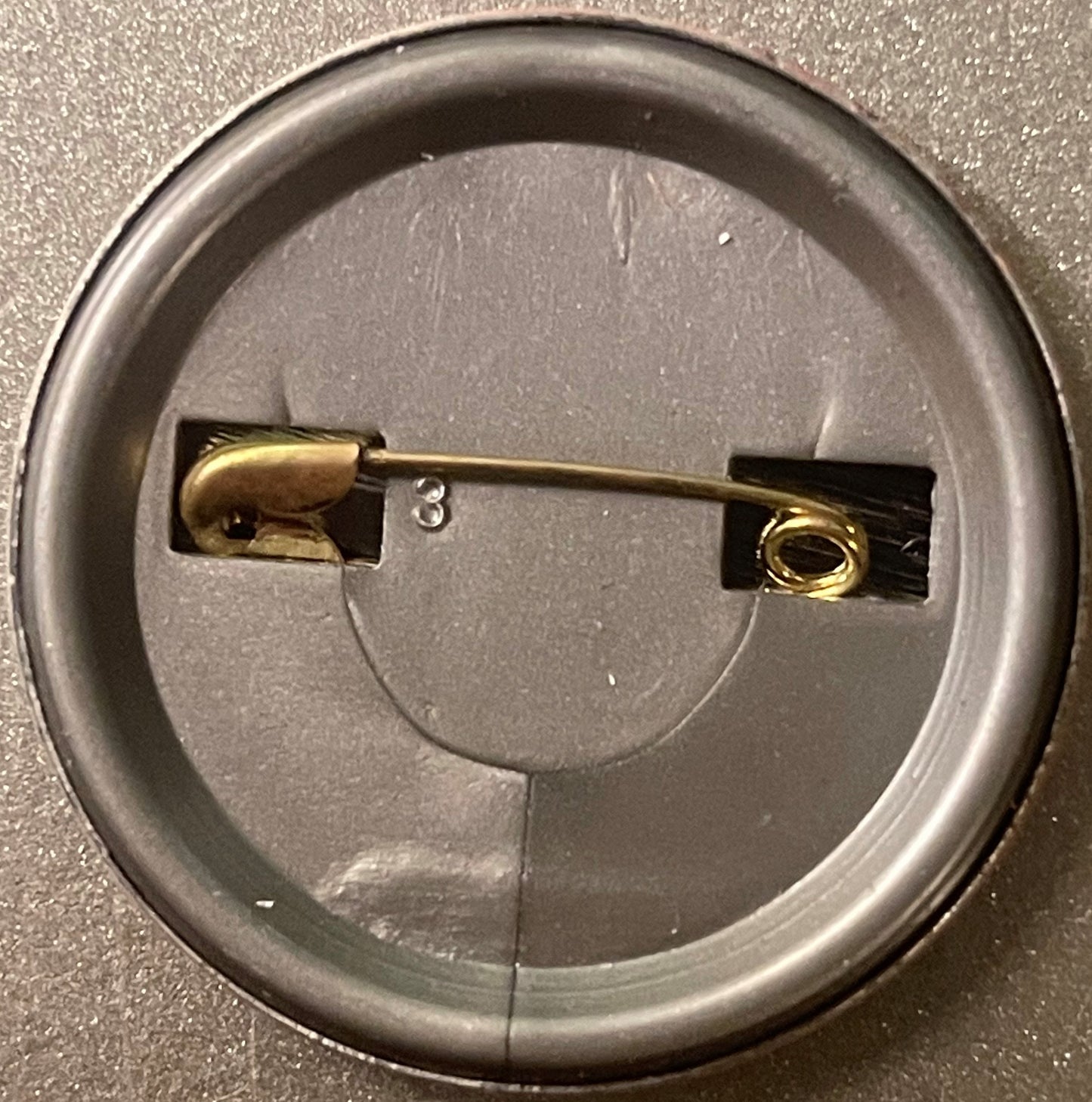 Back of a round metal pinback button showing brass fastener and black backing plate on gray surface