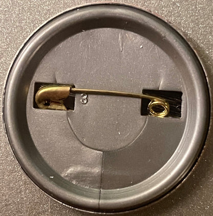 Back of a round metal pinback button showing brass fastener and black backing plate on gray surface