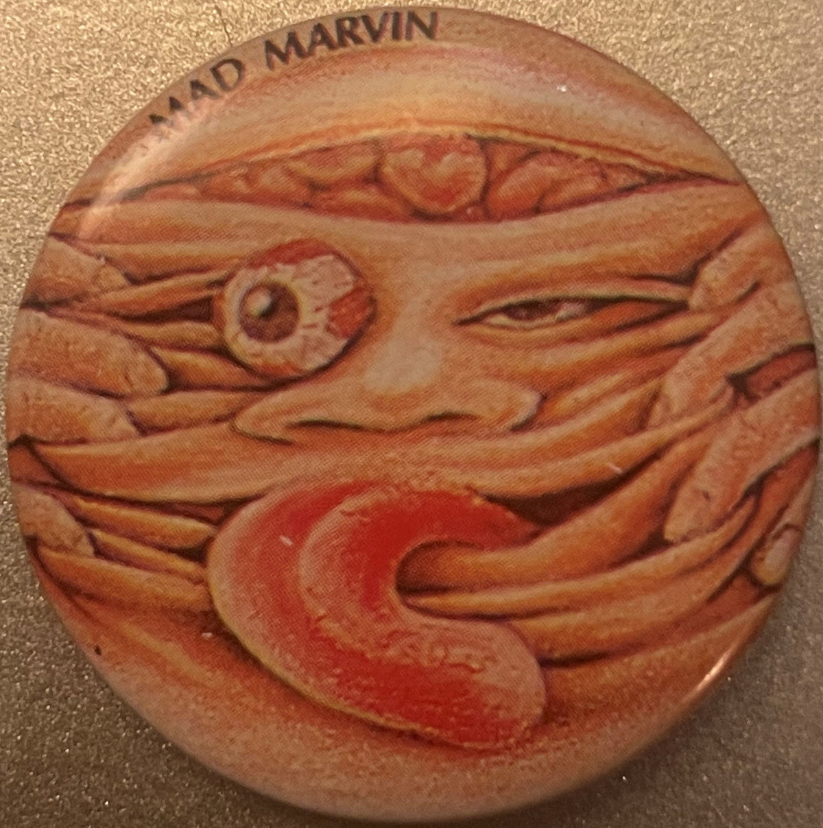 Circular clay-colored pinback button with embossed grotesque face featuring bulging eyes and protruding mouth with red paint 
