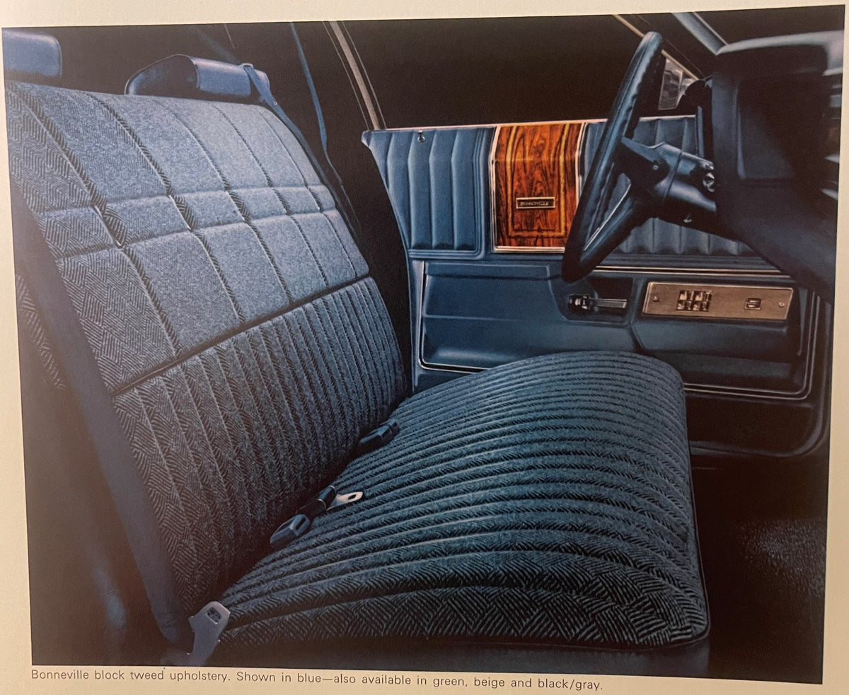 Interior view of 1974 Pontiac Bonneville showing blue block tweed upholstery on front bench seat and backrest, with wood-grain trim accent and steering wheel visible.