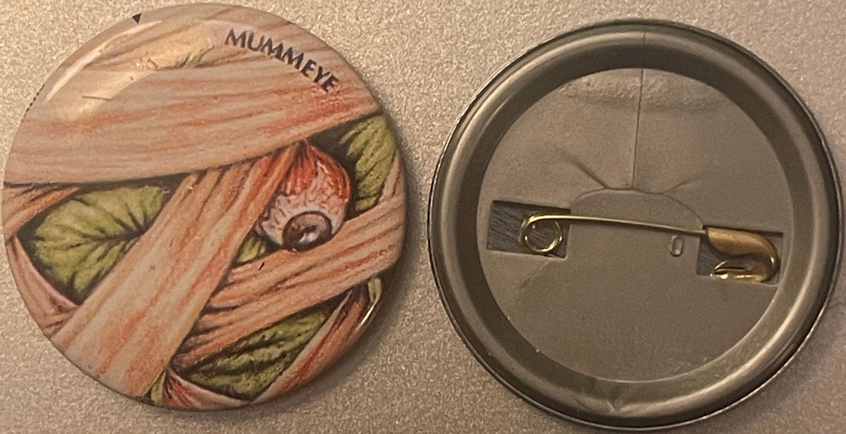 Front and back of a round pin badge featuring a mummified face wrapped in bandages with the word "MUMMEYE" printed at top