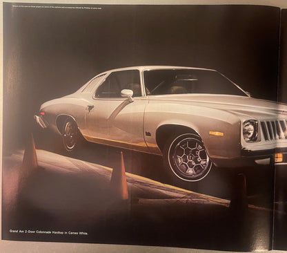 Cream-colored Pontiac Grand Am 2-door Colonnade Hardtop displayed against dark background with orange safety barriers visible