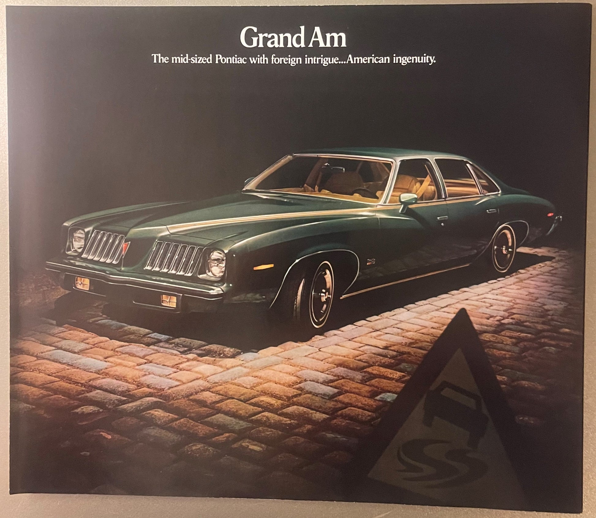 Dark green Pontiac Grand Am sedan with gold interior on brick surface, "Grand Am" title and tagline visible at top of brochur
