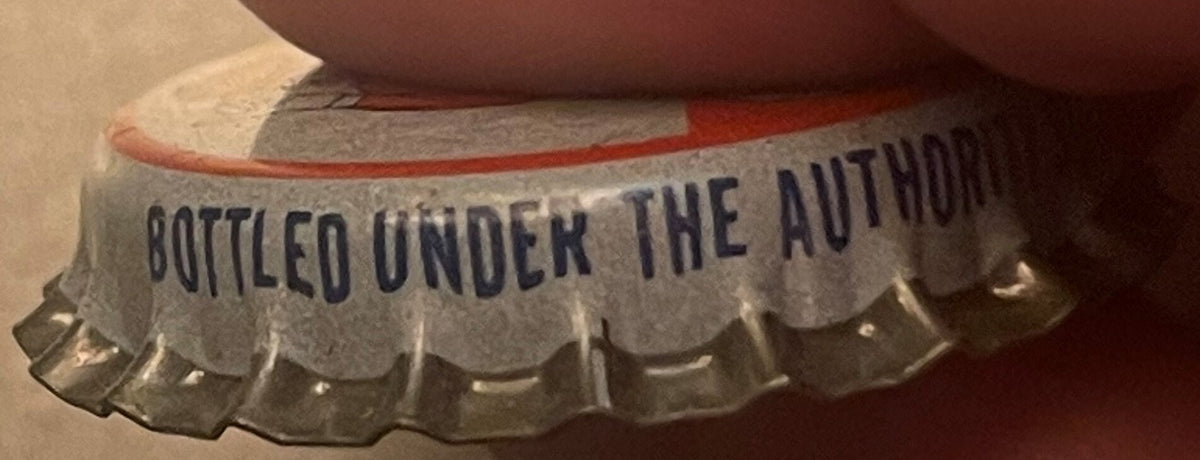 Close-up of vintage cork-lined soda bottle cap with scalloped metal edges, text reading "BOTTLED UNDER THE AUTHORITY" visible on crown, held by person's thumb for scale.