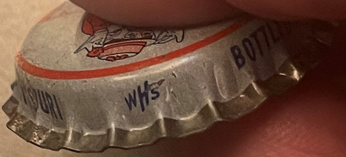 Side of a bottle cap held under a finger, showing part of the clown artwork, a red stripe, small blue 'WHS' marking, and 'BOTTL...' text.
