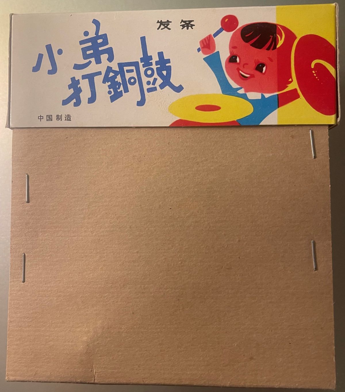 Vintage tin toy drummer box with red character and blue text on cardboard, shown closed from above with plain brown box