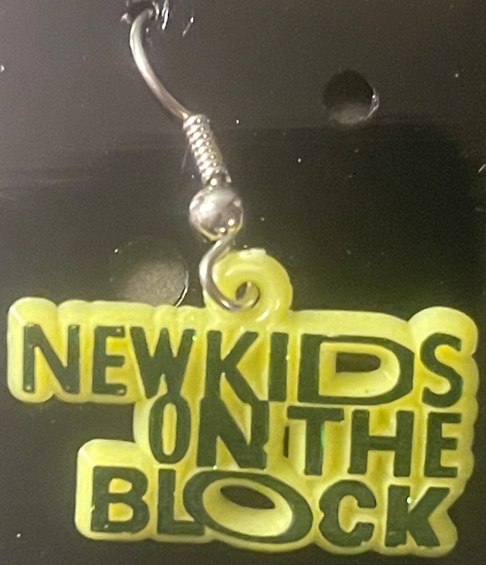 Yellow plastic charm earring with silver hook displayed against dark background, text reads "NEW KIDS ON THE BLOCK