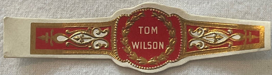 Ornate gold and red cigar band with "TOM WILSON" text in center, decorative scrollwork on sides, photographed flat against te