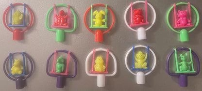 Ten colorful plastic animal swing pencil toppers from the 1980s arranged in two rows on a gray surface. Each features a small animal figure inside a swing frame with a cylindrical handle in assorted bright colors including red, green, pink, purple, white, and yellow.