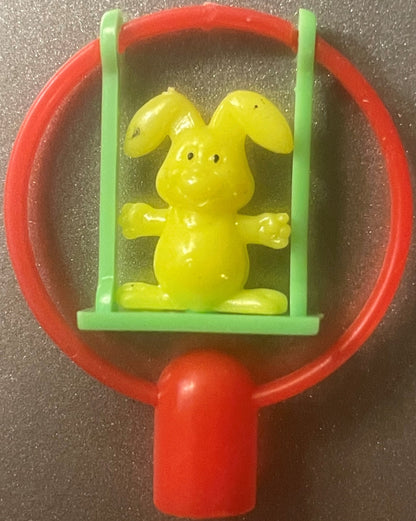A yellow bunny pencil topper with floppy ears on a green swing inside a red frame