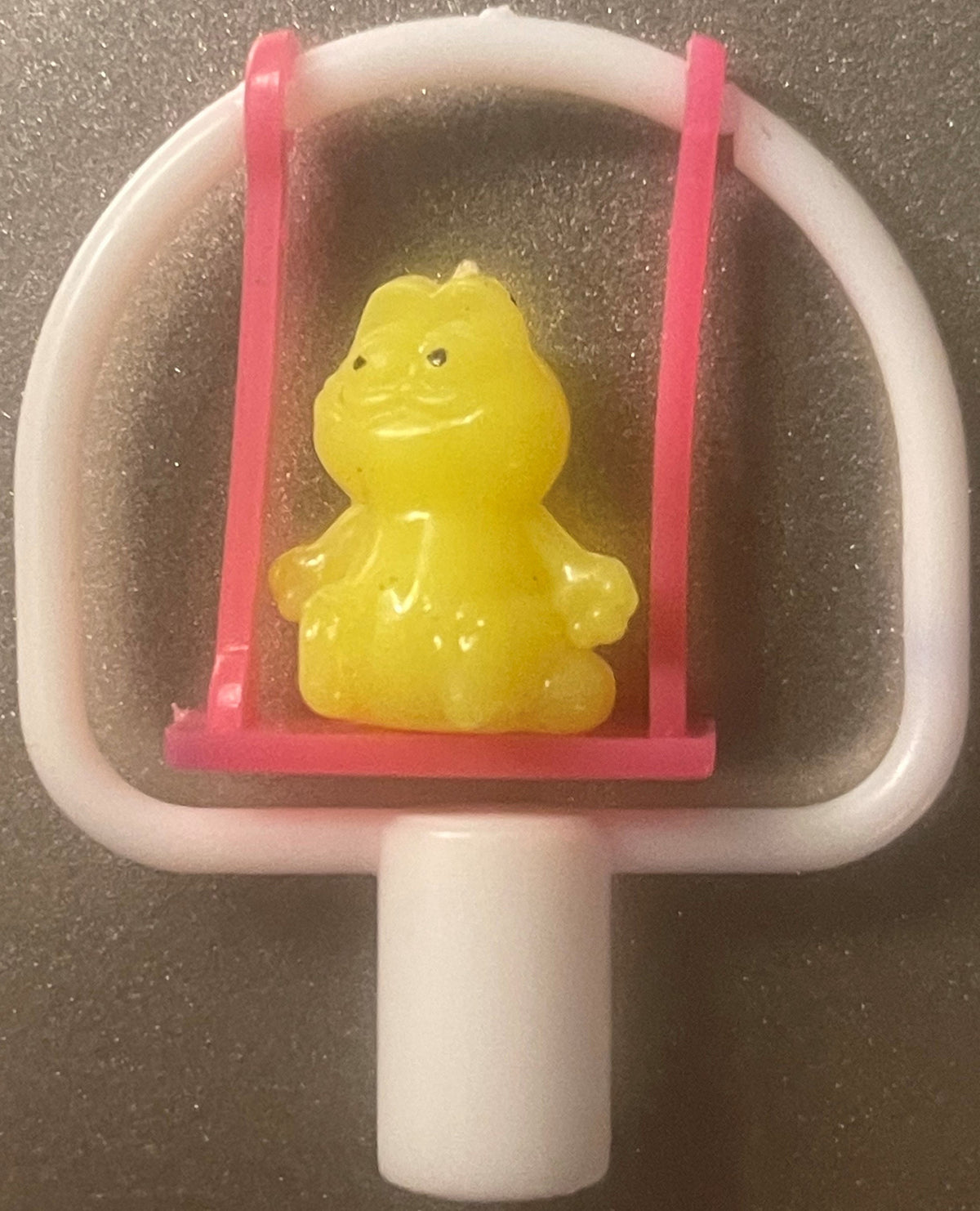 A yellow cat pencil topper on a pink swing inside a white frame