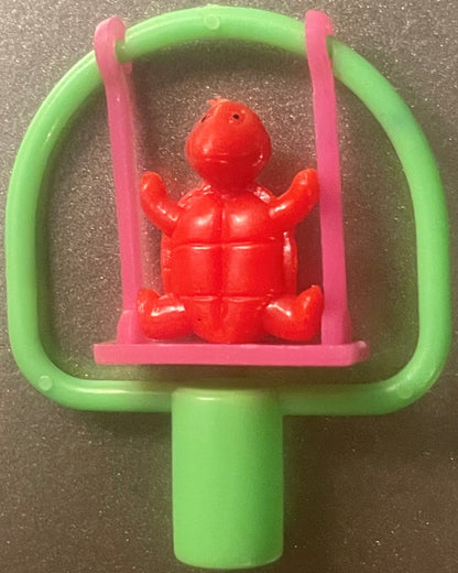A red turtle pencil topper, arms raised, on a pink swing inside a green frame