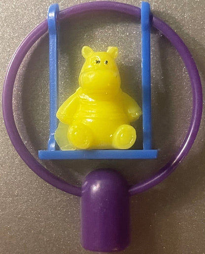 A yellow hippo pencil topper on a blue swing inside a purple frame