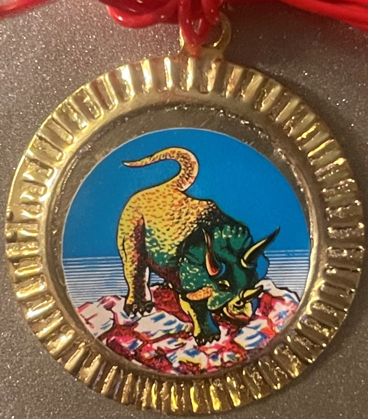 Circular brass charm with scalloped edge featuring colorful dinosaur illustration: yellow spotted creature with green body