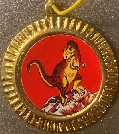 Gold-rimmed charm pendant with red enamel background featuring a yellow and brown dinosaur illustration, displayed against a 