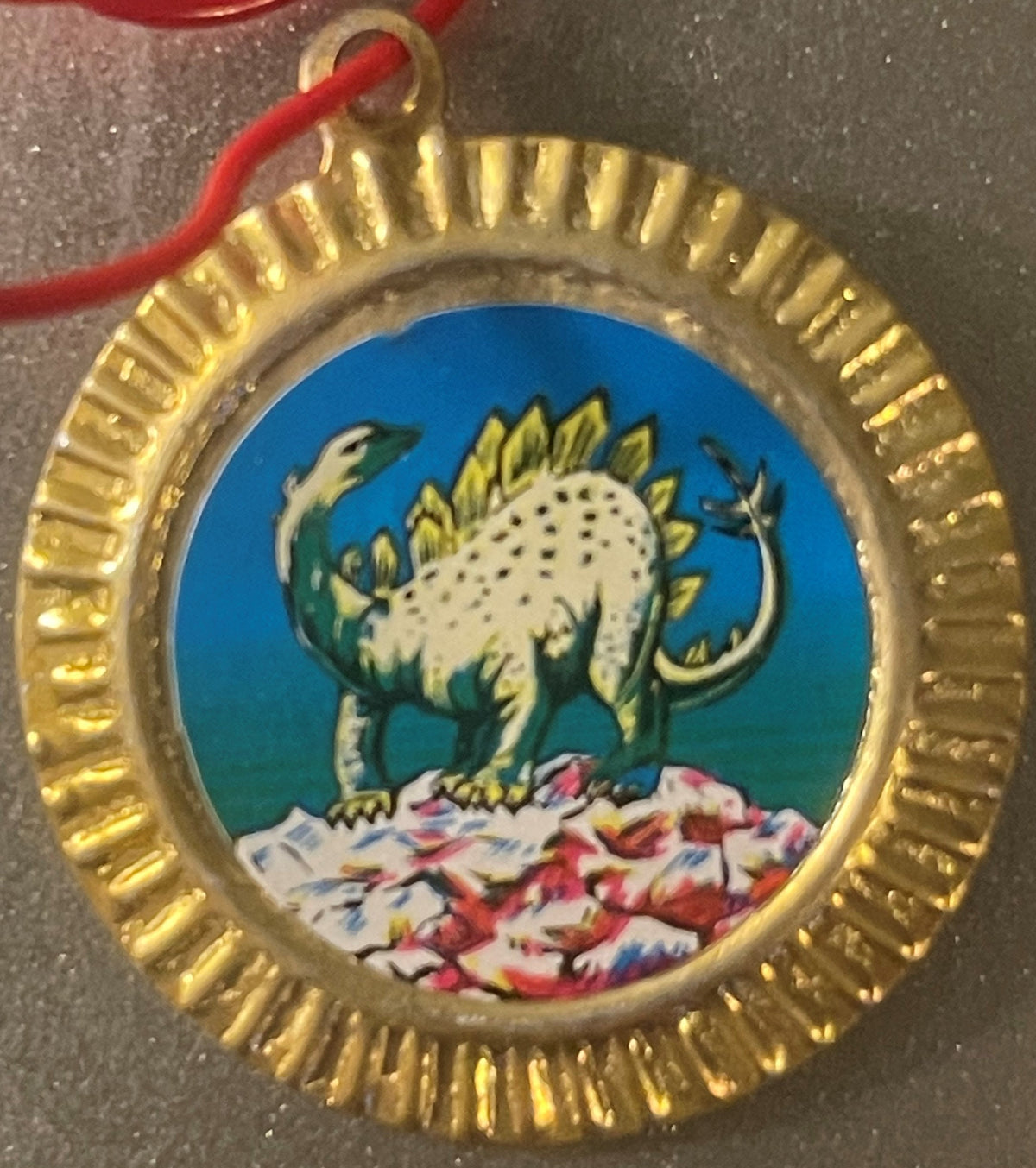Vintage tin dinosaur charm with gold ridged border, featuring a white stegosaurus with yellow plates on blue background and c