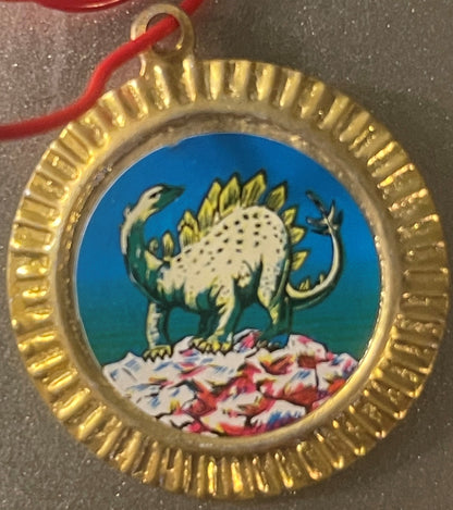 Vintage tin dinosaur charm with gold ridged border, featuring a white stegosaurus with yellow plates on blue background and c