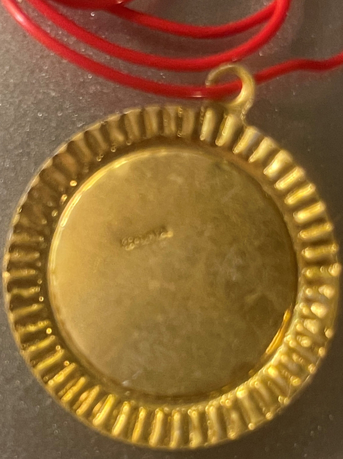 Gold-colored oval pendant with ribbed border on red cord, shown close-up against gray surface