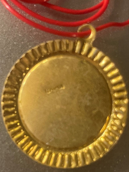 Gold-colored oval pendant with ribbed border on red cord, shown close-up against gray surface