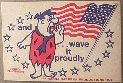 Vintage 1975 Fred Flintstone bicentennial sticker with American flag, stars, and text "and...wave it proudly" on cream backgr