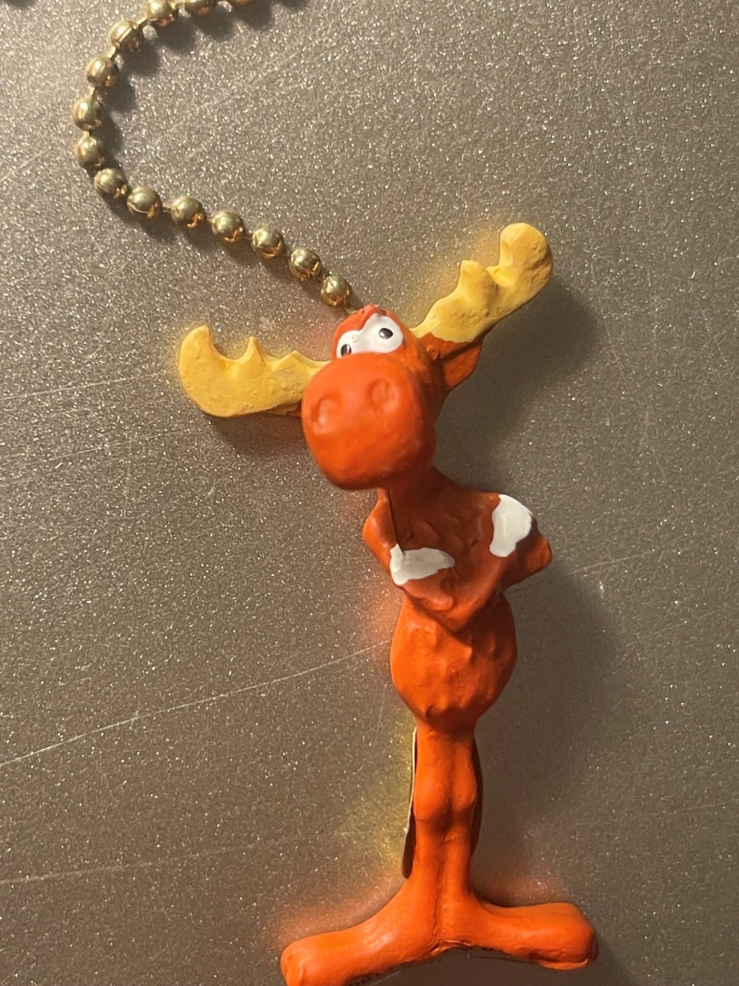 Vintage orange moose ceiling fan pull with yellow antlers and white chest details, attached to gold bead chain on gray textur