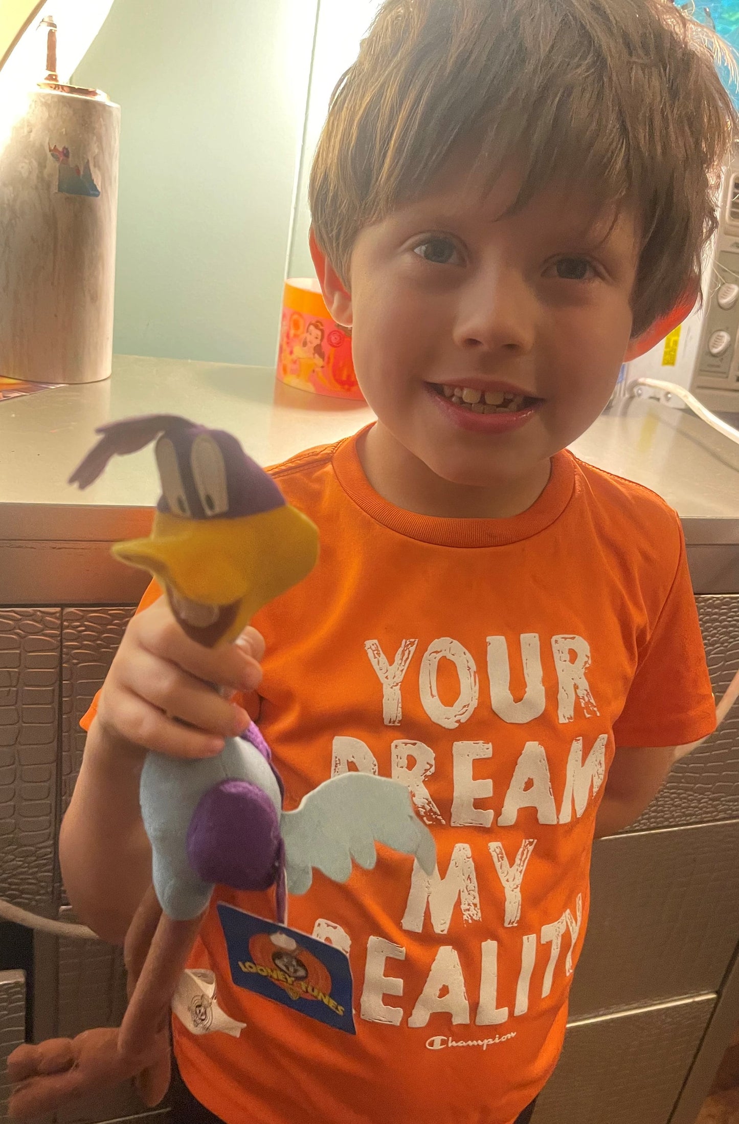 Young child in orange shirt holding vintage Road Runner plush toy with purple and yellow fabric, showing tag detail