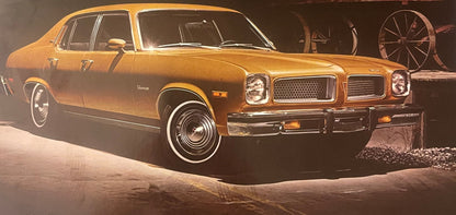 Golden-brown 1974 Pontiac Ventura sedan shown in three-quarter view against rustic barn setting with wagon wheel