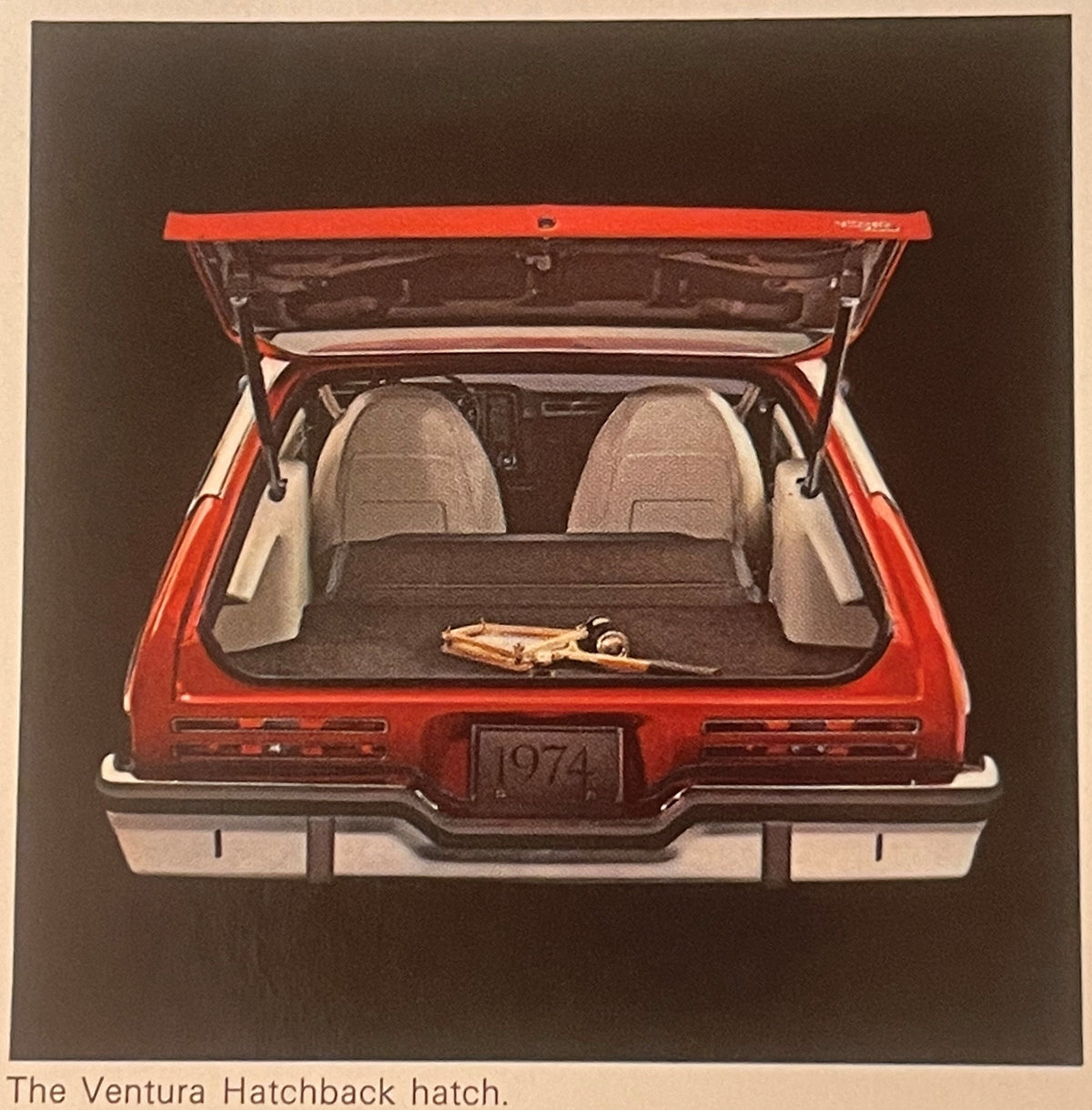 Rear view of a red and white 1974 Pontiac Ventura with open hatchback showing interior seats and cargo area against brown