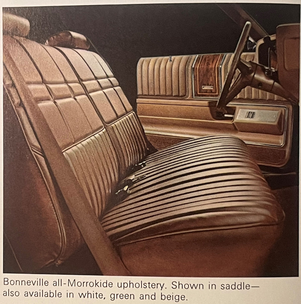 1974 Pontiac Bonneville interior showing tan Morrokide upholstery with quilted backrest and horizontal-ribbed bench seat, steering wheel visible on right, caption notes saddle color also available in white, green, and beige.