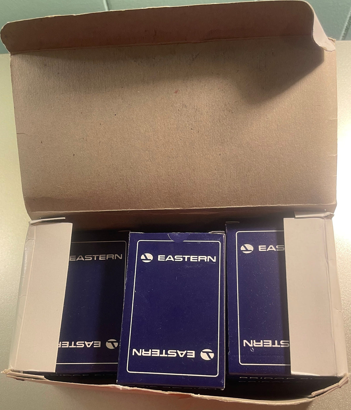 Overhead view of open cardboard box containing three sealed decks of Eastern Air Lines playing cards with navy blue boxes