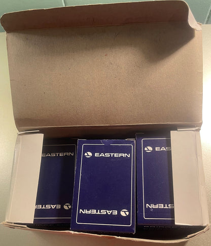 Overhead view of open cardboard box containing three sealed decks of Eastern Air Lines playing cards with navy blue boxes