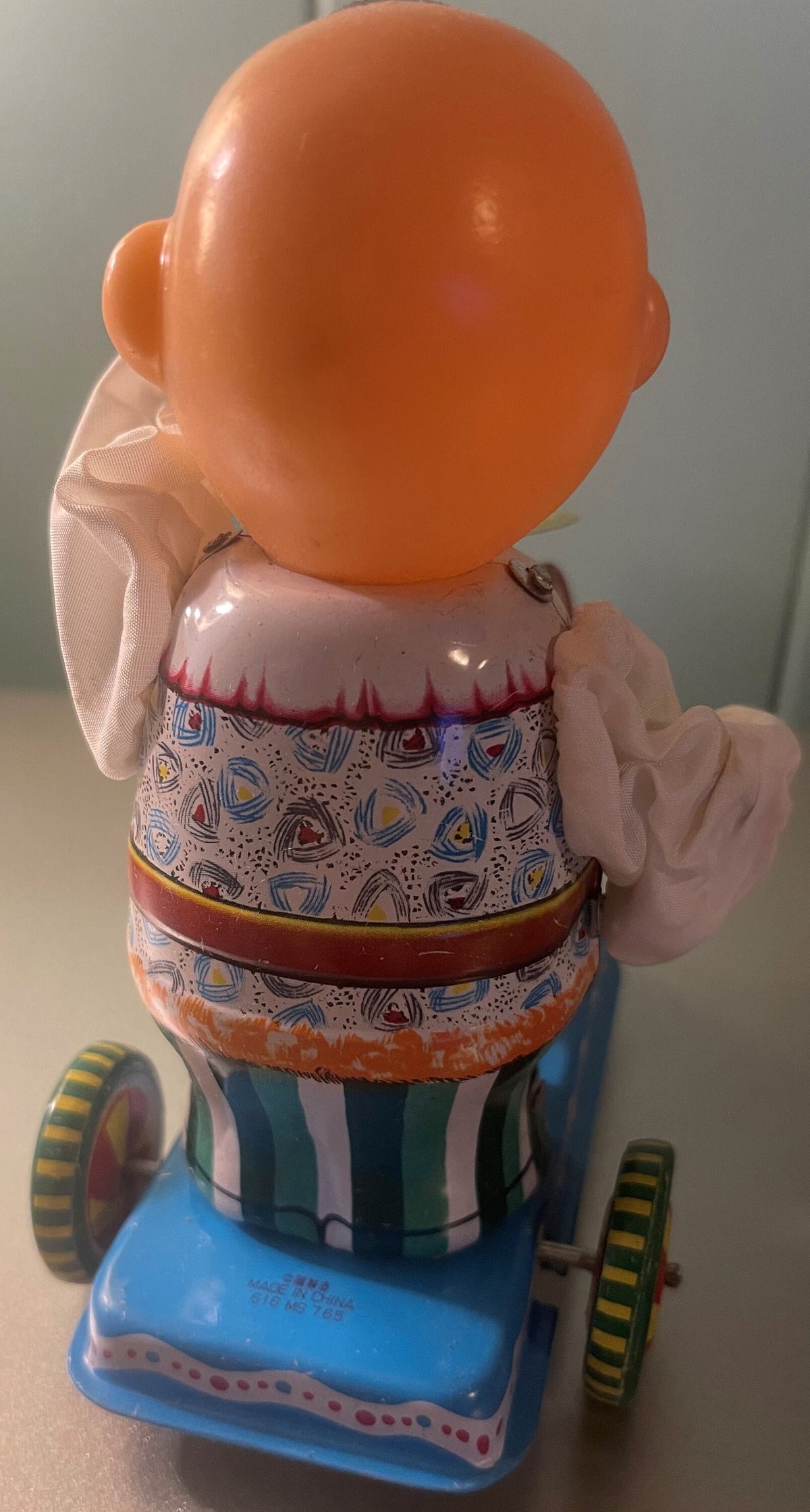Rear view of vintage tin wind-up toy drummer with orange head, patterned shirt, striped pants, blue base