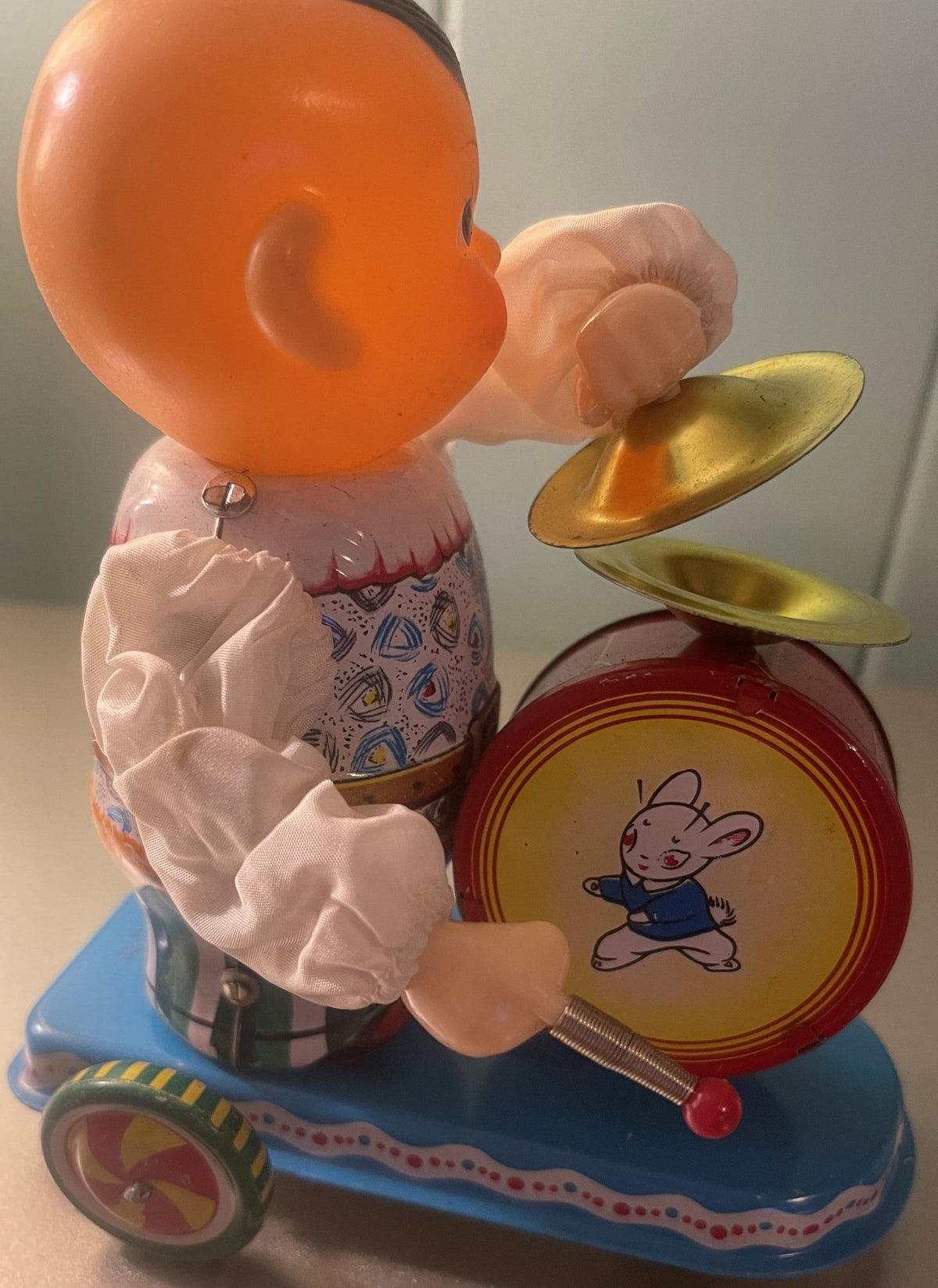 Vintage clockwork tin drummer toy with orange head, pink body, gold cymbals, and red drum with bunny illustration, mounted