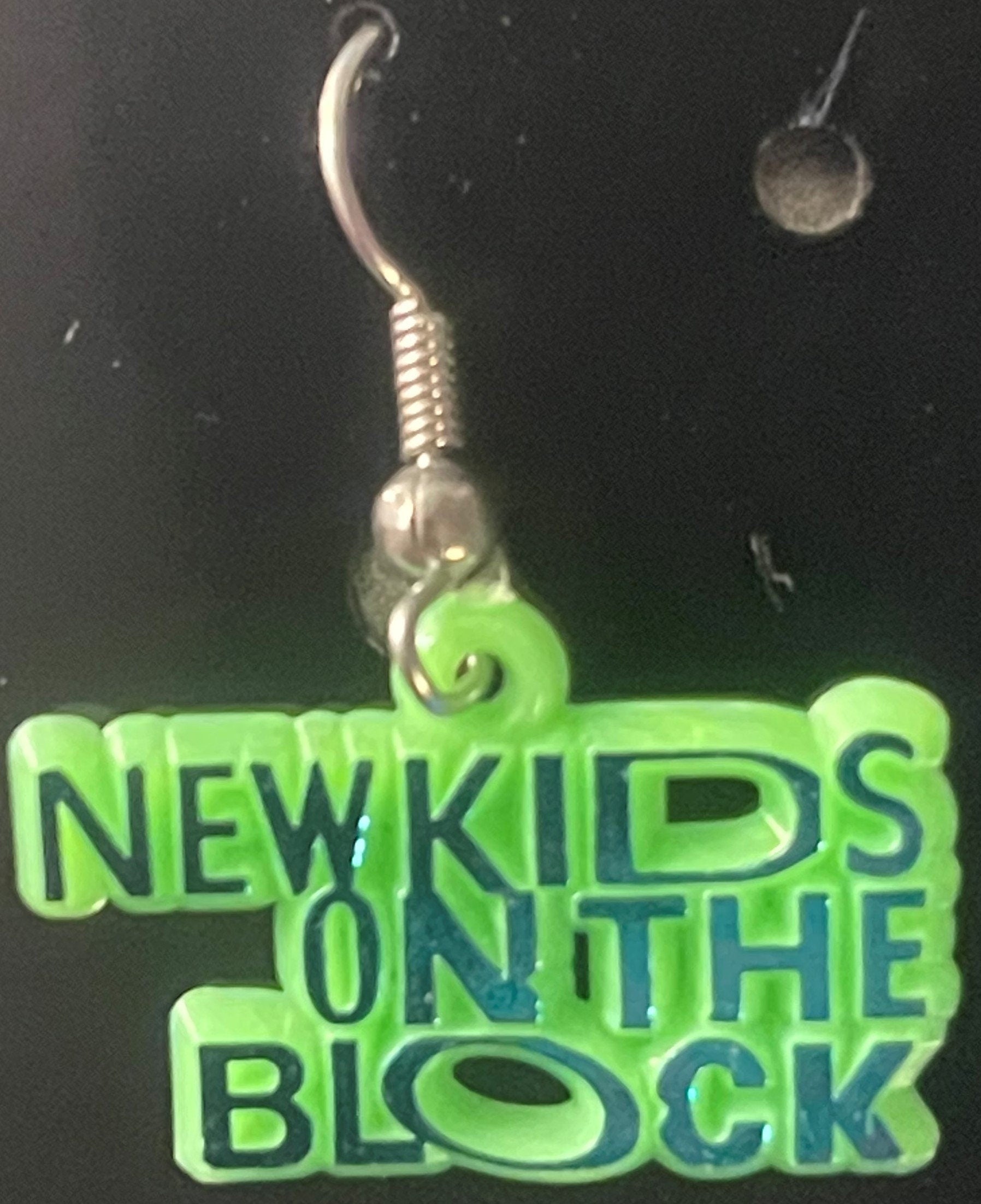 Neon green NKOTB earring charm with silver hook and coiled stem, displayed on matching green text card against dark backgroun