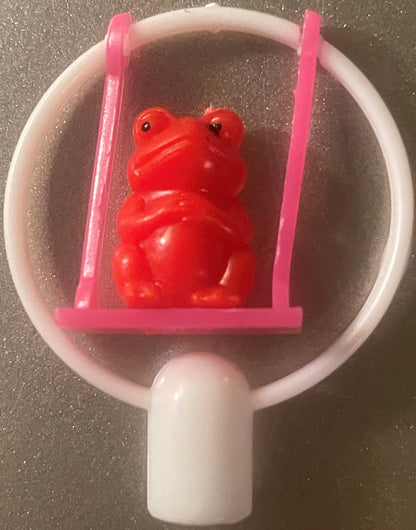 A red frog pencil topper on a pink swing inside a white frame