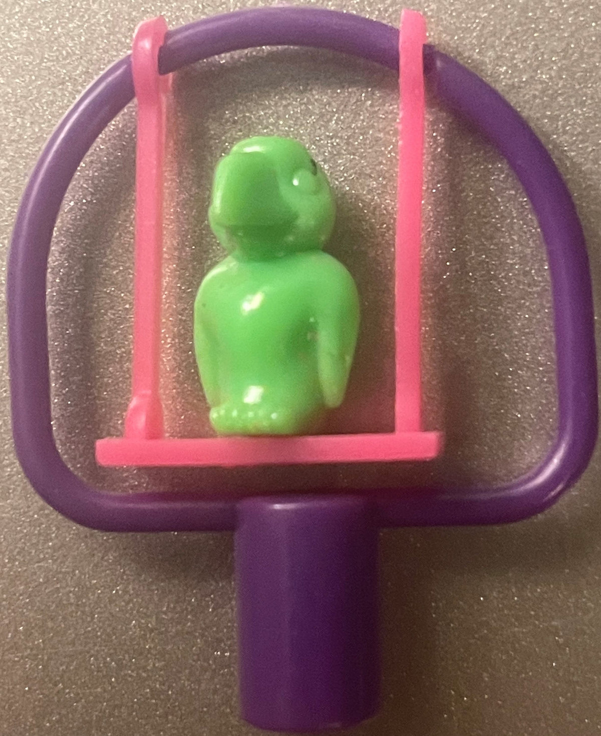 Green plastic bird figurine seated on pink swing suspended from purple frame with handles, vintage 1980s pencil topper displayed against textured surface.