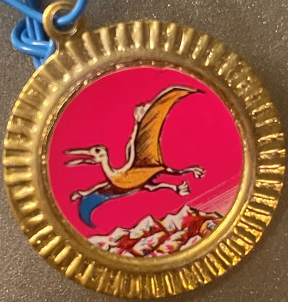 Vintage tin dinosaur charm with gold scalloped border, featuring a brown pterodactyl in flight on bright red background with 