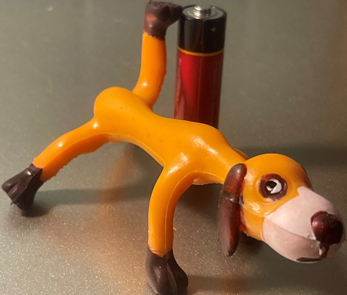 Vintage orange and brown bendable dog toy figure in stretched pose, with red and black battery-operated device visible in background for scale.