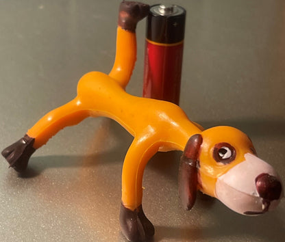 Vintage orange and brown bendable dog toy figure in stretched pose, with red and black battery-operated device visible in background for scale.