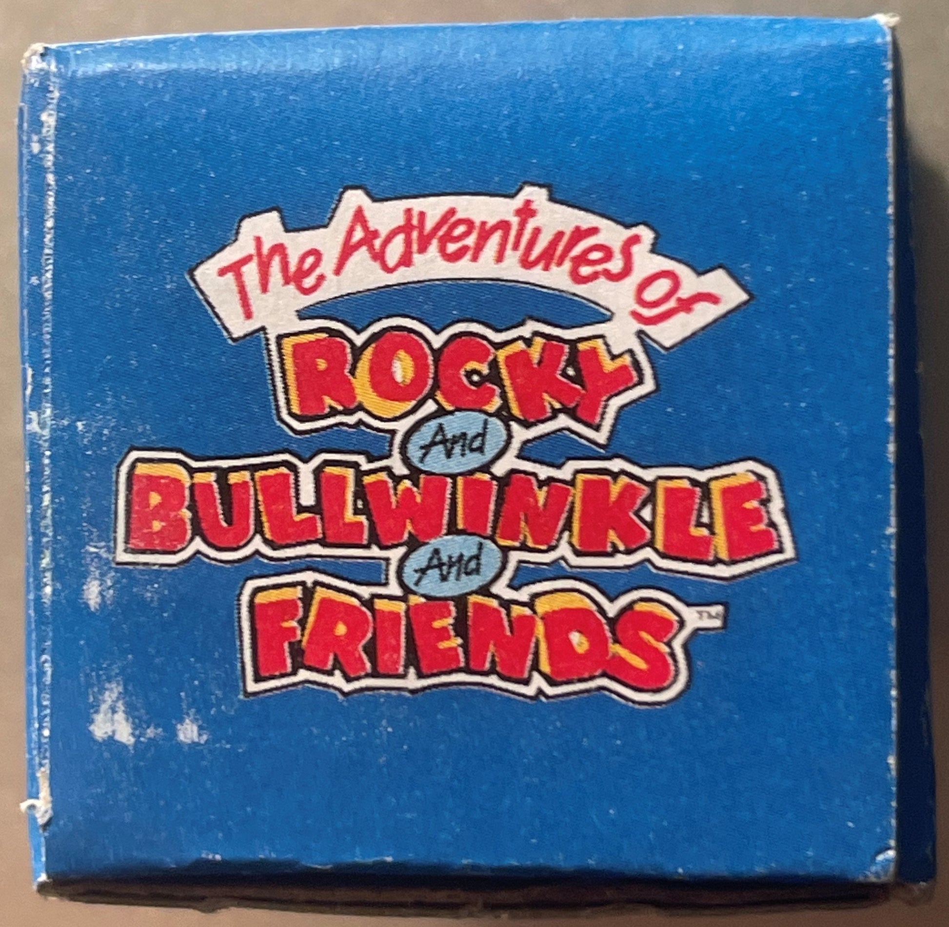 Blue box with red and yellow "The Adventures of Rocky and Bullwinkle and Friends" logo text, shown front-facing against tan b