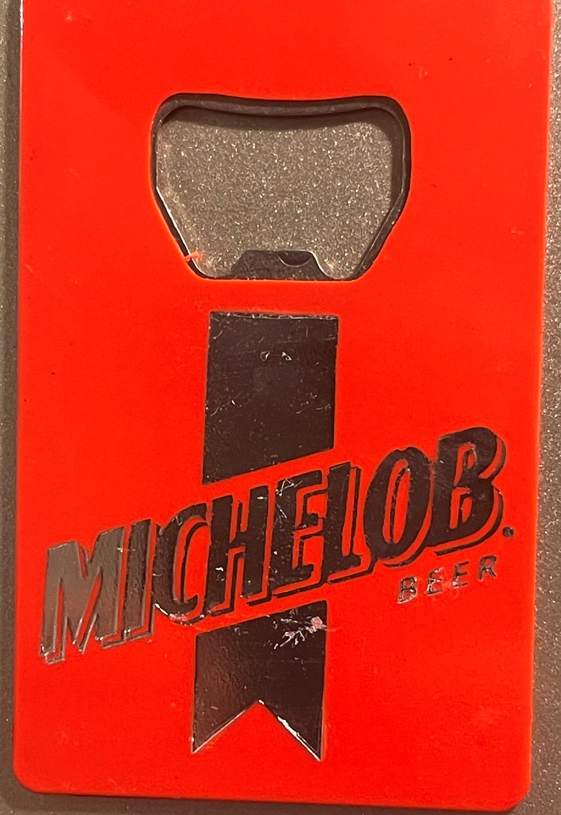 Red flat wallet-style bottle opener with embossed Michelob Beer logo and metal opener pad, shown against textured background