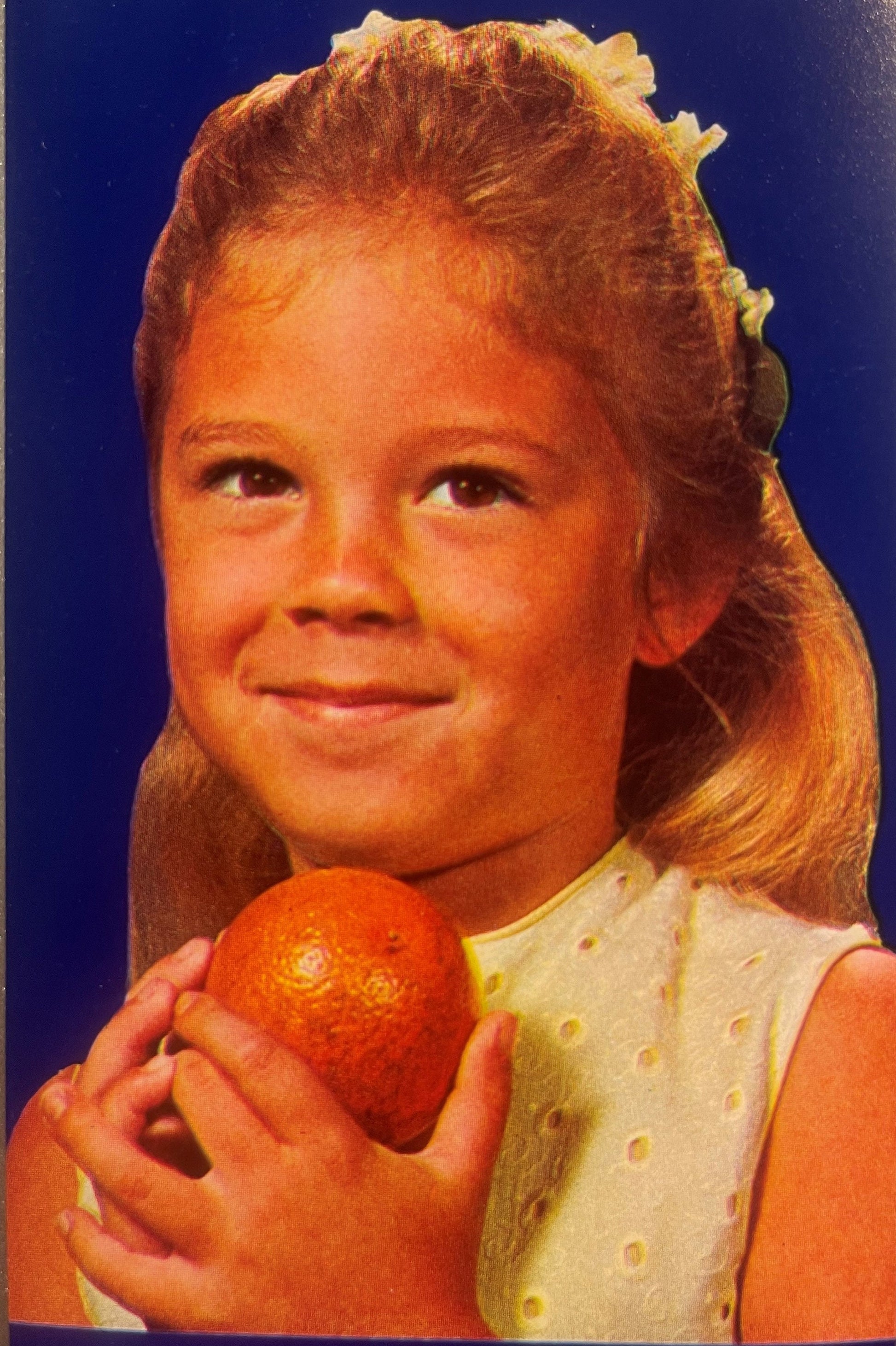 Young girl with reddish-blonde hair holding a red orange against dark blue background, wearing yellow and white dotted dress