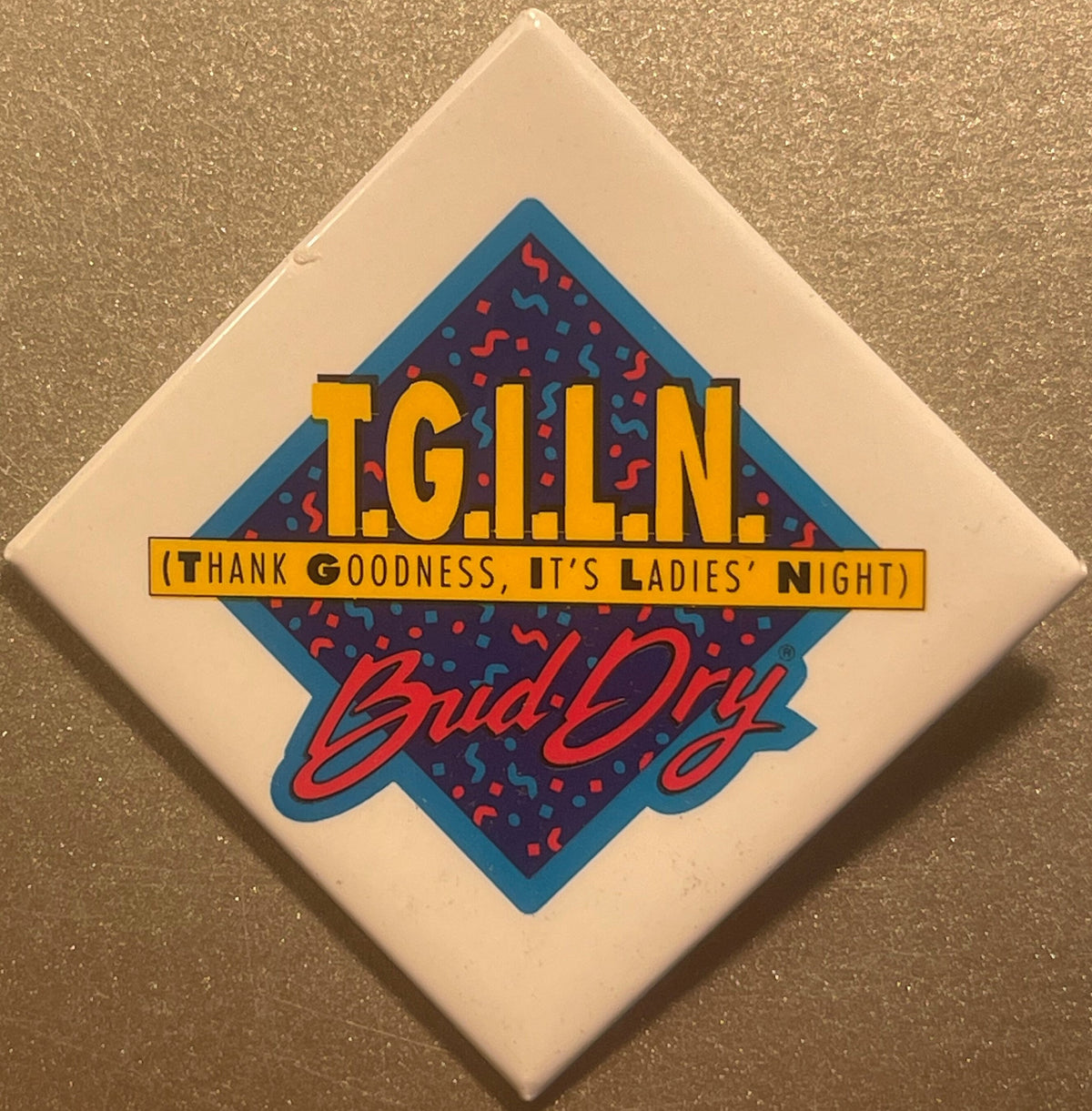 Square cream-colored pin with blue diamond shape featuring red and blue confetti pattern, gold "TGILN" text, yellow banner