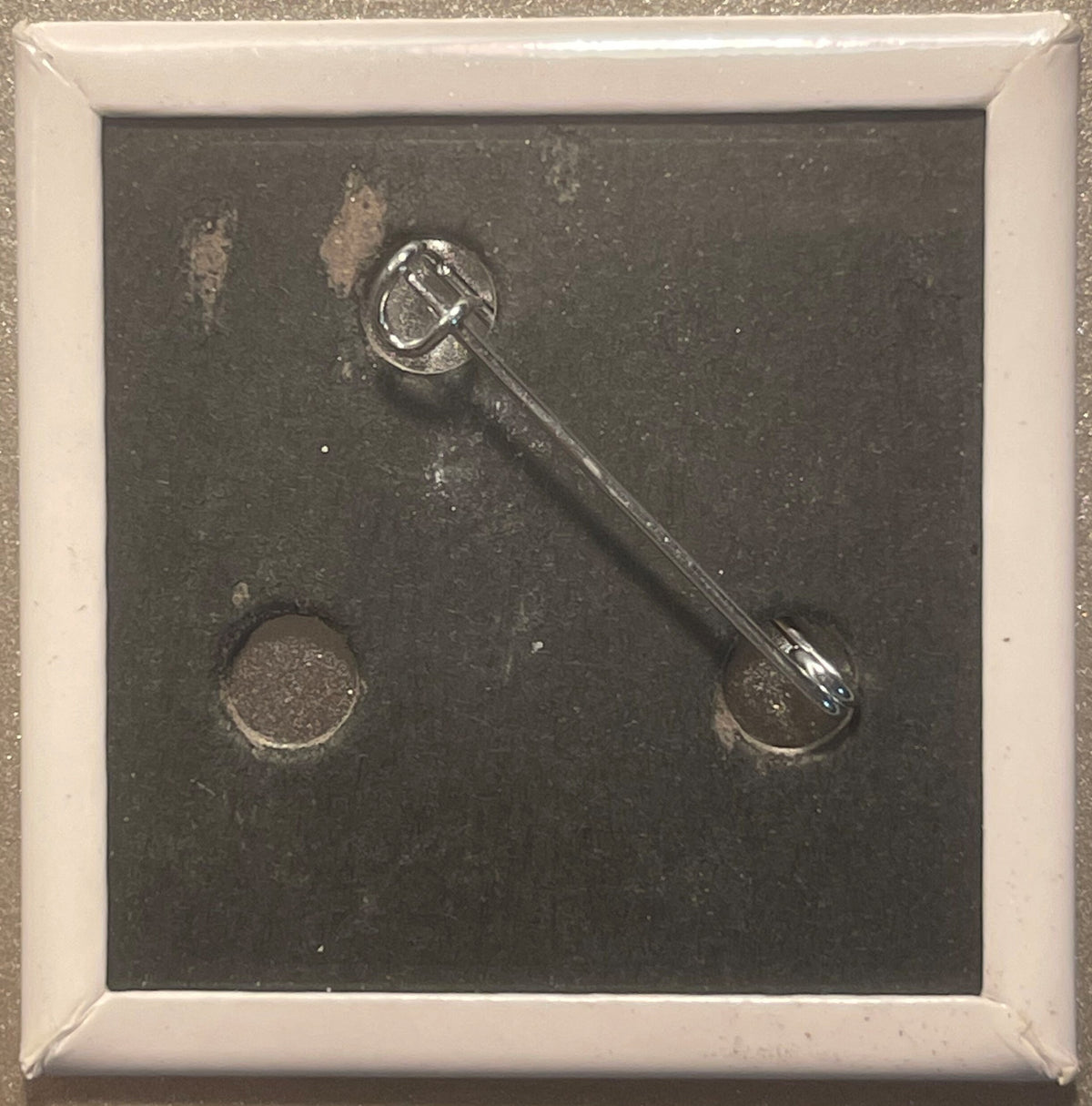 Silver-toned pin with circular top featuring crossed utensils design and spoon-shaped bottom, displayed in white-framed case 